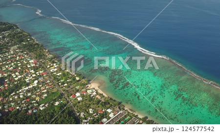 Turquoise ocean waves crashing against a vibrant coral reef contrast beautifully with lush green vegetation and colorful houses on a tropical island, captured from above. Exotic travel vacation 124577542