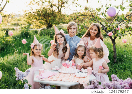 Summer outdoor kids birthday party. Group of happy Children celebrating birthday in park. Children blow candles on birthday cake. Kids party pink pastel decoration and food. Presents and sweets. 124579247