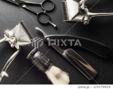 Various vintage barber tools including clippers, scissors, a straight razor, and a shaving brush are arranged on a dark surface, representing classic grooming equipment. Various vintage barber tools including clippers, scissors, a straight razor, and a shaving brush are arranged on a dark surface, representing classic grooming equipment. 124579929
