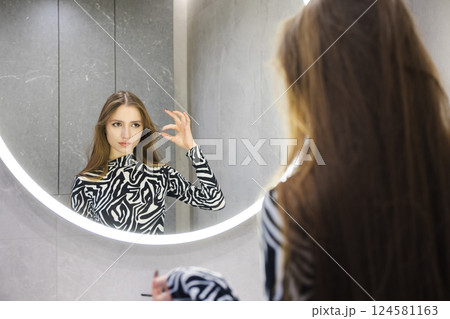 Teenage girl looking into a round illuminated mirror while styling her hair in a modern bathroom with a calm, focused expression 124581163