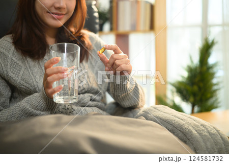 Smiling woman holding fish oil capsule and a glass of water sitting in a living room 124581732