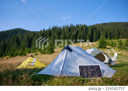 Three tents, including yellow one with striped chair, set up in grassy field with solar panel. Backdrop features lush forest and rolling hills under clear blue sky, creating tranquil camping scene. 124582757