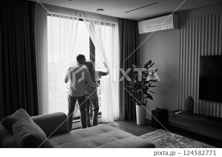Young couple looking at view from their new apartment. Happy couple hugging. Back view of male and his girlfriend next to big window at home. Black and white image. Young couple looking at view from their new apartment. Happy couple hugging. Back view of male and his girlfriend next to big window at home. Black and white image. 124582771