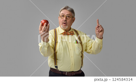 Senior teacher holding red apple and raising finger, looking surprised against grey studio background. 124584314