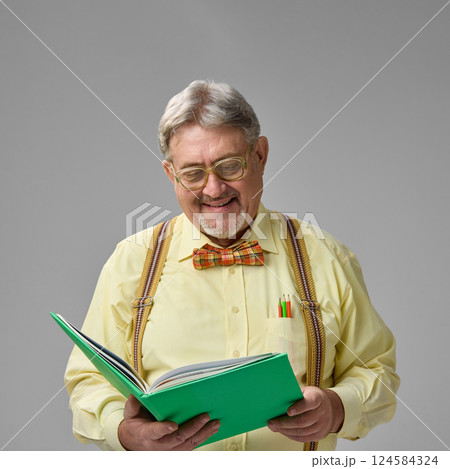 Elderly man smiling while reading book, enjoying academic material on neutral background. Calm moment, love for reading, daily work joy. 124584324