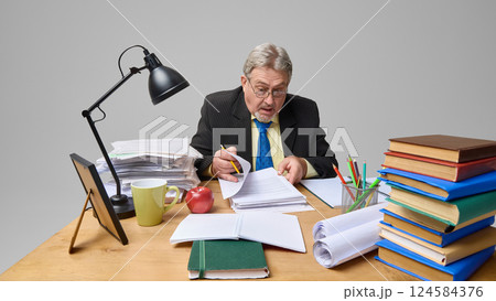 Shocked senior male professor looking at papers, sitting at wooden desk surrounded by books and folders 124584376