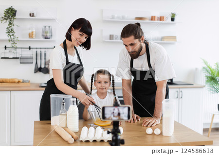 Caucasian family of bloggers filming cooking tutorial. Woman, man, and young girl preparing recipe, kneading dough in modern, bright kitchen environment while camera records. Caucasian family of bloggers filming cooking tutorial. Woman, man, and young girl preparing recipe, kneading dough in modern, bright kitchen environment while camera records. 124586618