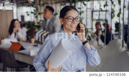 A Professional Woman in an Office Environment Engaging on the Phone with Detailed Blueprints 124590106