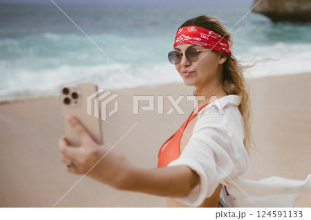 Beautiful happy woman doing selfie on beach summer. Young smiling girl making selfie on smartphone over sea background, cloudy weather. Romantic lady with laughing emotional face make photo on phone 124591133