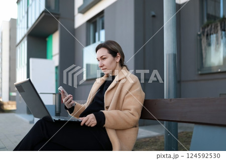 Young woman using laptop and smartphone while sitting on a bench outside a modern building in the 124592530