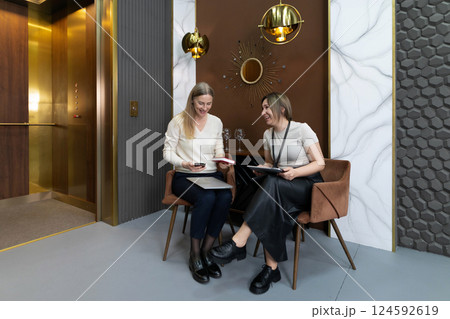 Two women engaged in conversation while sitting in a stylish lounge area near an elevator in a 124592619