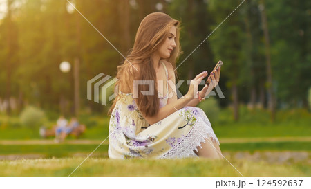 Young attractive woman in summer dress sits on green grass with mobile phone in hands. A park. Summer. Recreation. 124592637