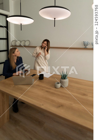 Two women discussing ideas in a modern office setting with warm lighting and minimalist decor 124593768