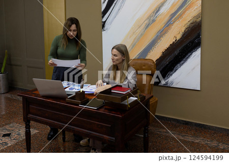 Two women collaborating at a modern office desk reviewing documents and discussing project details Two women collaborating at a modern office desk reviewing documents and discussing project details 124594199