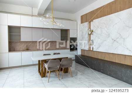 A modern, spacious kitchen with light facades, wooden trim, marble tiles, a table, upholstered chairs, an empty countertop, and an electric oven. 124595273