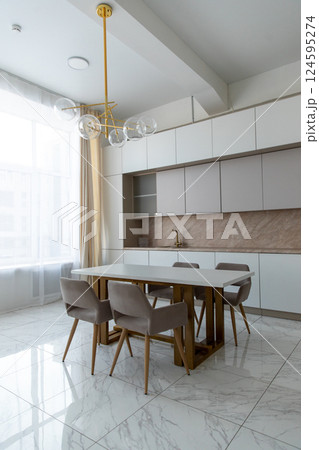A modern, spacious kitchen with light facades, wooden trim, marble tiles, a table, upholstered chairs, an empty countertop. Minimalistic design. 124595274