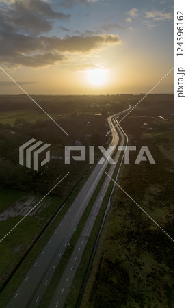 A video captured during golden hour, showing a winding road through a rural landscape, fields with water reflections, and distant buildings, all under a warm, sunset-lit sky. 124596162