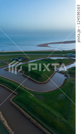 A drone video showing a unique landscape of man-made mounds and waterways near the sea, with a house on the grassy terrain, and a distant view of the coastline stretching under a cloudy sky. A drone video showing a unique landscape of man-made mounds and waterways near the sea, with a house on the grassy terrain, and a distant view of the coastline stretching under a cloudy sky. 124596163
