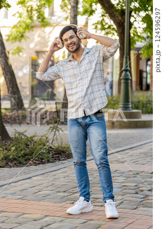 Portrait of happy young Indian man wearing wireless headphones listening to music on city street Portrait of happy young Indian man wearing wireless headphones listening to music on city street 124596297