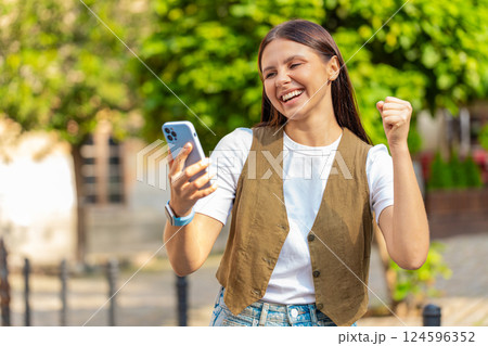 Happy Caucasian young woman using smartphone celebrating win good message news on city town street Happy Caucasian young woman using smartphone celebrating win good message news on city town street 124596352
