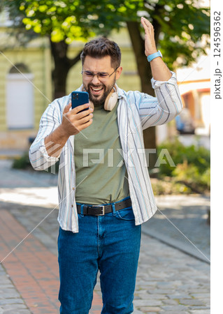 Upset young man using smartphone typing browsing, loses becoming shocked bad news on city street Upset young man using smartphone typing browsing, loses becoming shocked bad news on city street 124596362