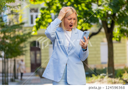 Caucasian mature businesswoman looking shocked while reading bad news on mobile phone on city street 124596368
