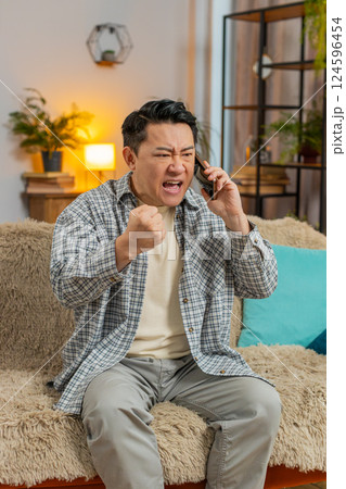 Angry young Chinese man arguing on smartphone phone while sitting on couch in living room at home 124596454