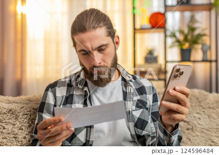 Young man sorting bills at home, sofa budget check, phone in hand, multiple papers, calm focus vibe Young man sorting bills at home, sofa budget check, phone in hand, multiple papers, calm focus vibe 124596484