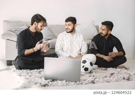 Indian male friends sitting on a floor while watching football match on laptop. Men emotionally disscussing. Men wearing black and white indian traditional clothes. Indian male friends sitting on a floor while watching football match on laptop. Men emotionally disscussing. Men wearing black and white indian traditional clothes. 124604522