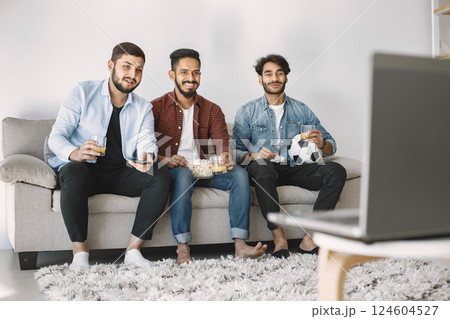 Three young excited male friends sitting on a coach while watching football match on laptop. Men drinking juice and eating popcorn. Men wearing shirts and t-shirts. 124604527