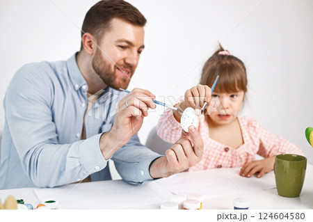 Portrait of a charming girl with Down syndrome wearing rose shirt and her bearded father. Girl sitting at desk in the kitchen painting Easter colored eggs. Girl and her father preparing Easter Portrait of a charming girl with Down syndrome wearing rose shirt and her bearded father. Girl sitting at desk in the kitchen painting Easter colored eggs. Girl and her father preparing Easter 124604580