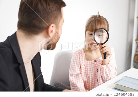 Male teacher and girl with Down syndrome sitting at desk. Girl holding a magnifying glass. Education for disabled children concept. Male teacher and girl with Down syndrome sitting at desk. Girl holding a magnifying glass. Education for disabled children concept. 124604588