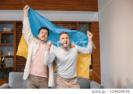 Male friends gathered at home to watch a football match. Two men standing and holding a big flag of Ukraine in hands. Men actively support their favorite team and feeling satisfied. Male friends gathered at home to watch a football match. Two men standing and holding a big flag of Ukraine in hands. Men actively support their favorite team and feeling satisfied. 124604636