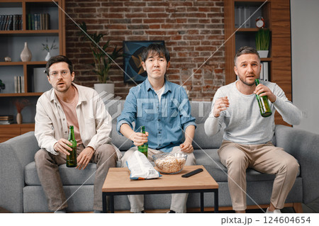 Front view of male friends gathered at home to watch a football match. Men sitting on the couch in front of the big screen TV and eating snacks. Men actively support their favorite team and feeling 124604654