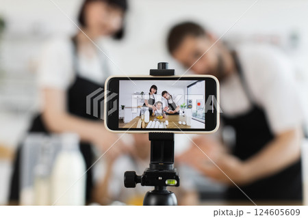 Caucasian adult parents and young child recording culinary activity, kneading dough in kitchen. Smartphone on tripod captures footage, representing blogging or vlogging, family cooking, and technology Caucasian adult parents and young child recording culinary activity, kneading dough in kitchen. Smartphone on tripod captures footage, representing blogging or vlogging, family cooking, and technology 124605609