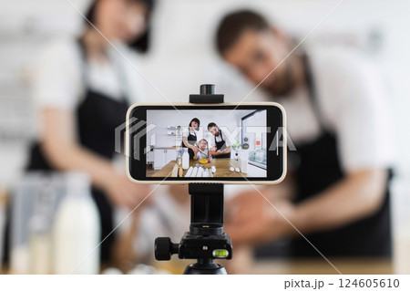 Caucasian family blogging cooking lesson using tripod-mounted smartphone. Adults assist child making dough in glass bowl Caucasian family blogging cooking lesson using tripod-mounted smartphone. Adults assist child making dough in glass bowl 124605610