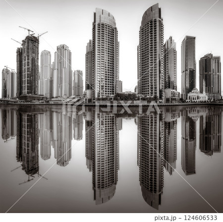 Monochrome city skyline with tall skyscrapers reflecting on calm water under a cloudy sky, Dubai Marina bay UAE 124606533