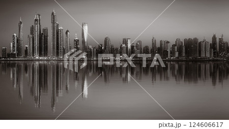 Monochrome city skyline with tall skyscrapers reflecting on calm water under a cloudy sky, Dubai Marina bay UAE 124606617
