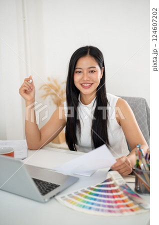 An asian woman is smiling at camera while holding a documents about design color on her working desk 124607202