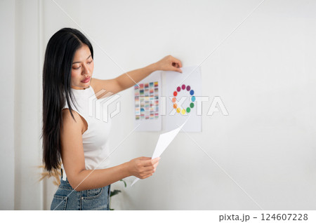 Asian woman is looking at the document in her hand while sticking printed color wheel chart on wall. 124607228