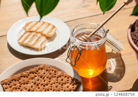Honey in a Jar With a Dipper Next to Toasted Bread on a Wooden Table, Sunlight Streaming Through Leaves a Delightful Breakfast 124608750