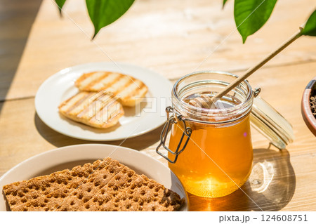 Honey Jar With Dipper and Grilled Bread on a Wooden Table, Sunlight Through Green Leaves, Cozy Cafe Vibe Honey Jar With Dipper and Grilled Bread on a Wooden Table, Sunlight Through Green Leaves, Cozy Cafe Vibe 124608751