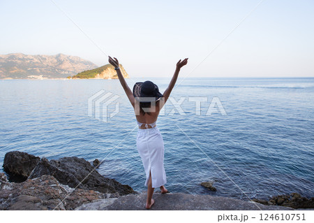 A woman in a white dress and sun hat joyfully raises her arms, overlooking a serene ocean and mountain landscape 124610751