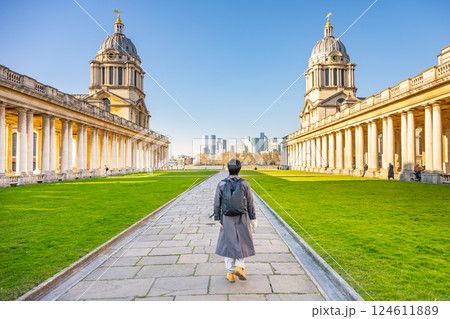 A visitor walks along the pathway amidst the stunning historical buildings of Greenwich Campus in London. With a clear blue sky, the city skyline can be spotted in the distance. 124611889