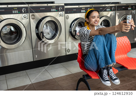 Housewife in laundromat taking selfie on mobile phone while using washing machine 124612910
