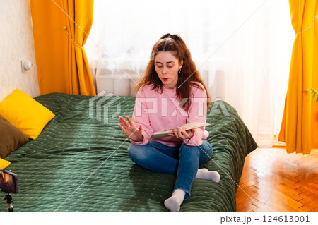 Female digital creator sitting on a cozy bed, recording a vlog with a smartphone on a tripod, sharing online business tips. Mid shot Female digital creator sitting on a cozy bed, recording a vlog with a smartphone on a tripod, sharing online business tips. Mid shot 124613001