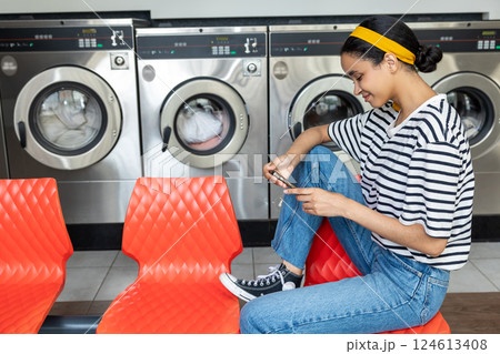 Woman with dark hair tapping and texting message on smartphone while sitting in public laundromat Woman with dark hair tapping and texting message on smartphone while sitting in public laundromat 124613408