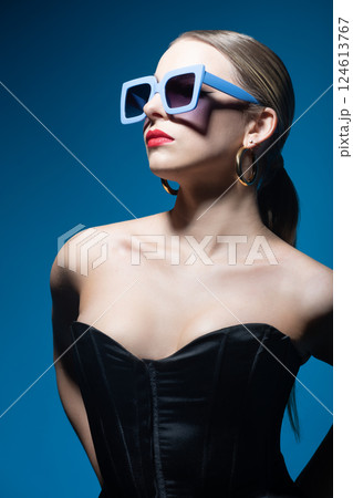 Woman portrait with classic hairstyle, big retro sunglasses, red lipstick,golden earrings and black corset in blue background 124613767