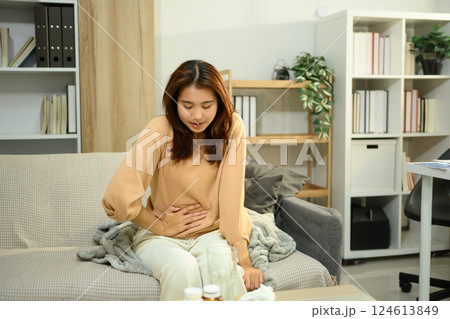 Asian woman suffering from stomach pain while sitting on sofa. Period cramps or gastrointestinal issues 124613849
