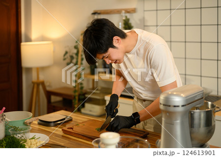Smiling young man chopping fresh parsley on wooden cutting board while preparing garlic butter in a cozy kitchen Smiling young man chopping fresh parsley on wooden cutting board while preparing garlic butter in a cozy kitchen 124613901